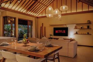 A modern living room with a dining area, a television displaying 'NETFLIX', and a large window with a view of greenery. - Apura Panoramic Villa 2BD Ubud (Ubud)