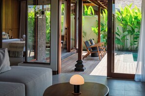 Quarto moderno com sofá, mesa com luminária, cama e vista para uma piscina e vegetação lá fora.