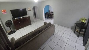Een woonkamer met een bank, televisie, ventilator, en een eethoek met stoelen en een tafel.