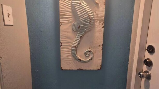 Een hal met een blauwe deur, een lamp erboven en een decoratie van een zeepaard aan de muur.