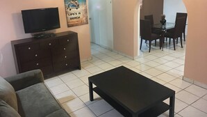Ein Wohnzimmer mit Fernseher, einer Couch, einem Couchtisch, einer dunklen Holzkommode und einem Essbereich mit Stühlen und einem Tisch.