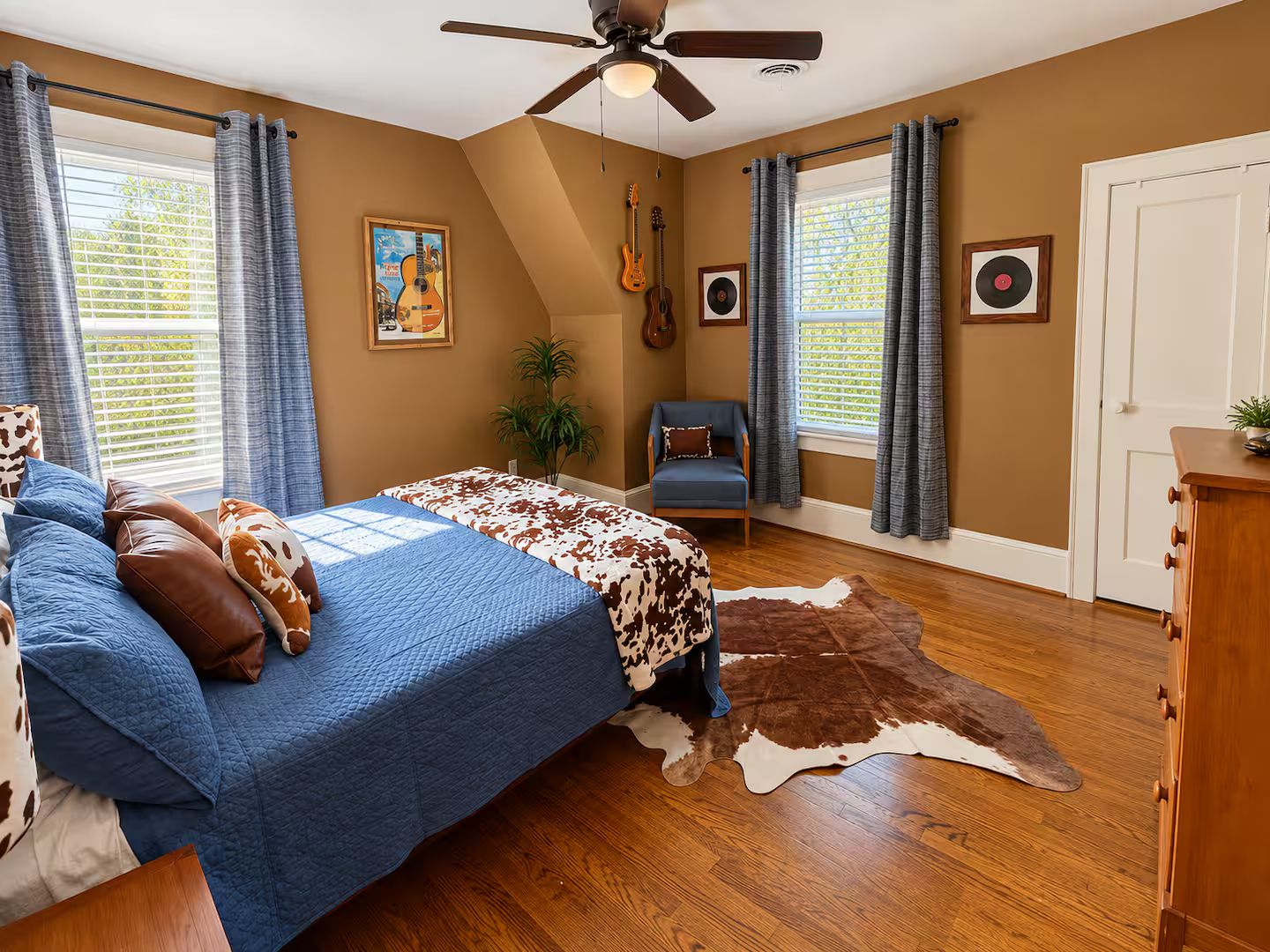 A bedroom with a bed, a ceiling fan, a guitar, a chair, a plant, and a door.