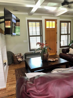 Ein Wohnzimmer mit Flachbildfernseher an der Wand, einem Couchtisch mit Dekoration, einem Ledersofa und einer Tür mit Jalousien.