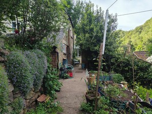 A narrow garden path leading to a house with a brick wall and a stone wall on one side, surrounded by various plants and gardening tools. - Cottage Sleeps 5 with Parking (Lydbrook)