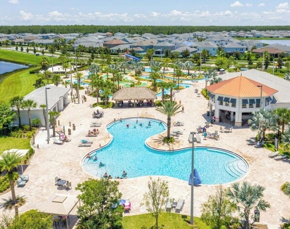 A swimming pool with lounge chairs and a thatched-roof pavilion, surrounded by palm trees and a view of a residential area. - 3180-302 Condo Resort Water Park Pools near Disney (Kissimmee)