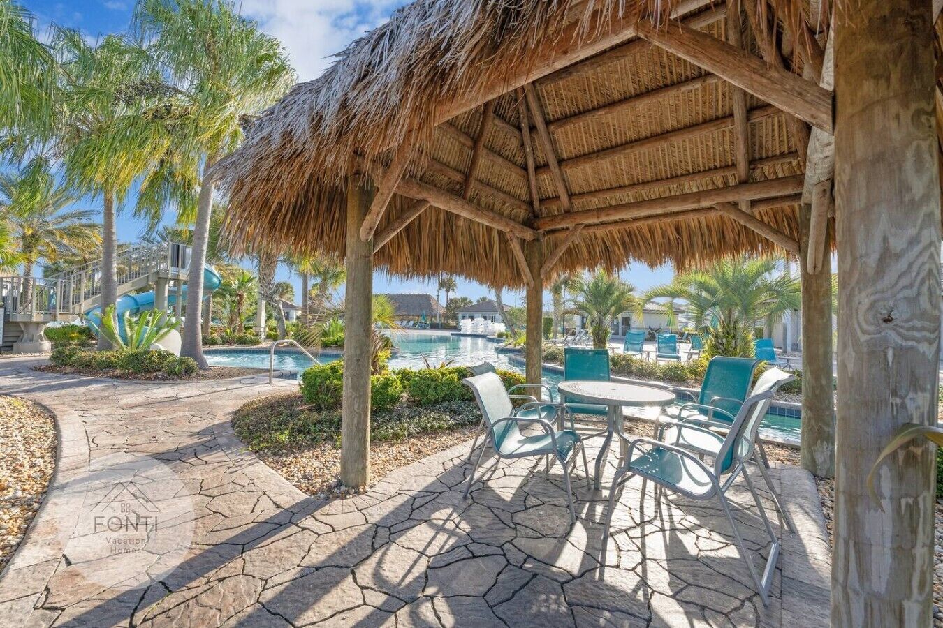 A covered outdoor seating area with a thatched roof, stone flooring, and a pool in the background.