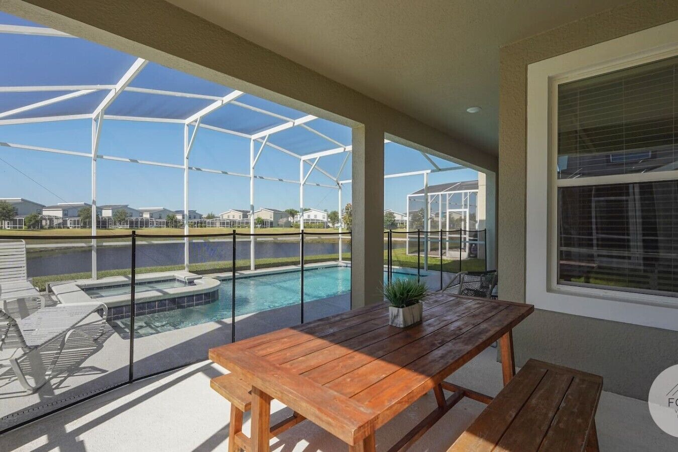 Ein überdachter Außenbereich mit Pool, Holztisch und Stühlen, mit Blick auf ein Gewässer und Wohnhäuser.
