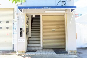 Entrada de um hotel com uma escadaria que leva ao interior, uma porta com persiana fechada e uma placa.