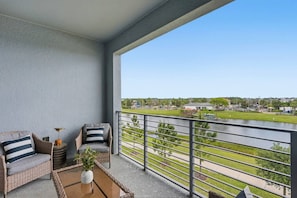Ein Balkon mit Korbmöbeln, einem kleinen Tisch mit Pflanze und Blick auf einen Fluss sowie entfernte Gebäude.