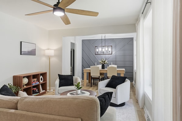 A modern living room with a ceiling fan, a dining area with a table and chairs, and a bookshelf.