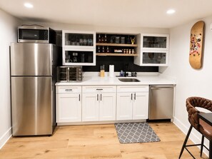 A modern kitchen with a stainless steel refrigerator, a microwave, a toaster, and a dishwasher. There are white cabinets with glass doors, a wooden shelf with glassware, and a patterned rug on the floor.