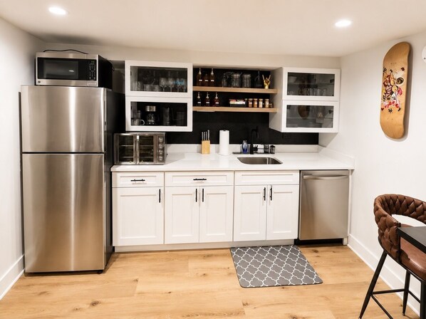 Una cocina moderna con un refrigerador de acero inoxidable, un microondas, un tostador y un lavavajillas. Hay armarios blancos con puertas de vidrio, un estante de madera con cristalería y una alfombra estampada en el suelo.