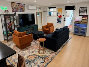 A living room with a TV, orange armchairs, a dark sofa, a patterned rug, and a bookshelf.