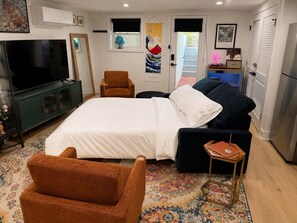 A bedroom with a bed, a TV, a chair, a sofa, a small table, and a door leading to a bathroom.