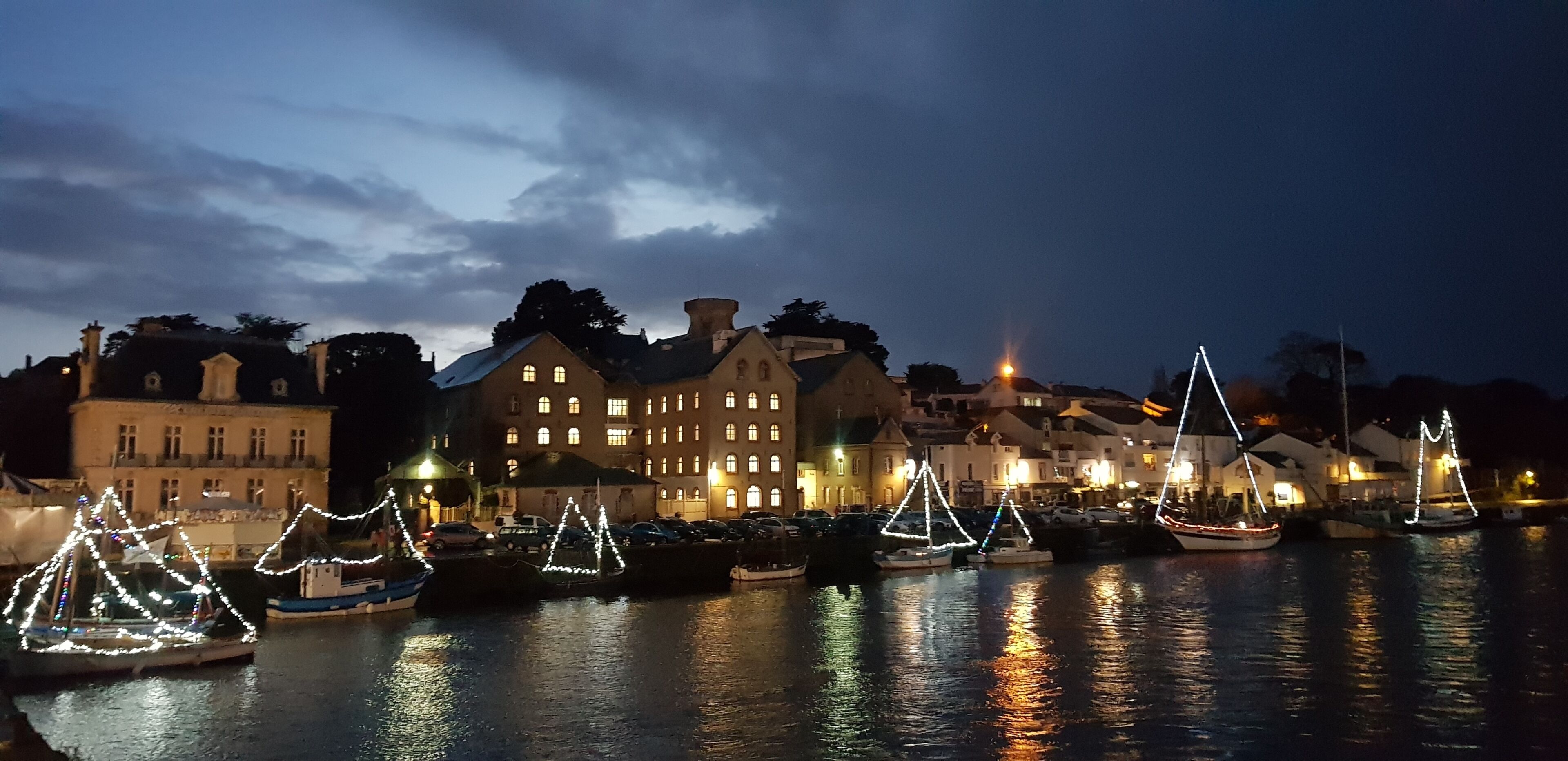 A waterfront with boats decorated with lights, a large building with multiple windows, and a calm water surface reflecting the lights.