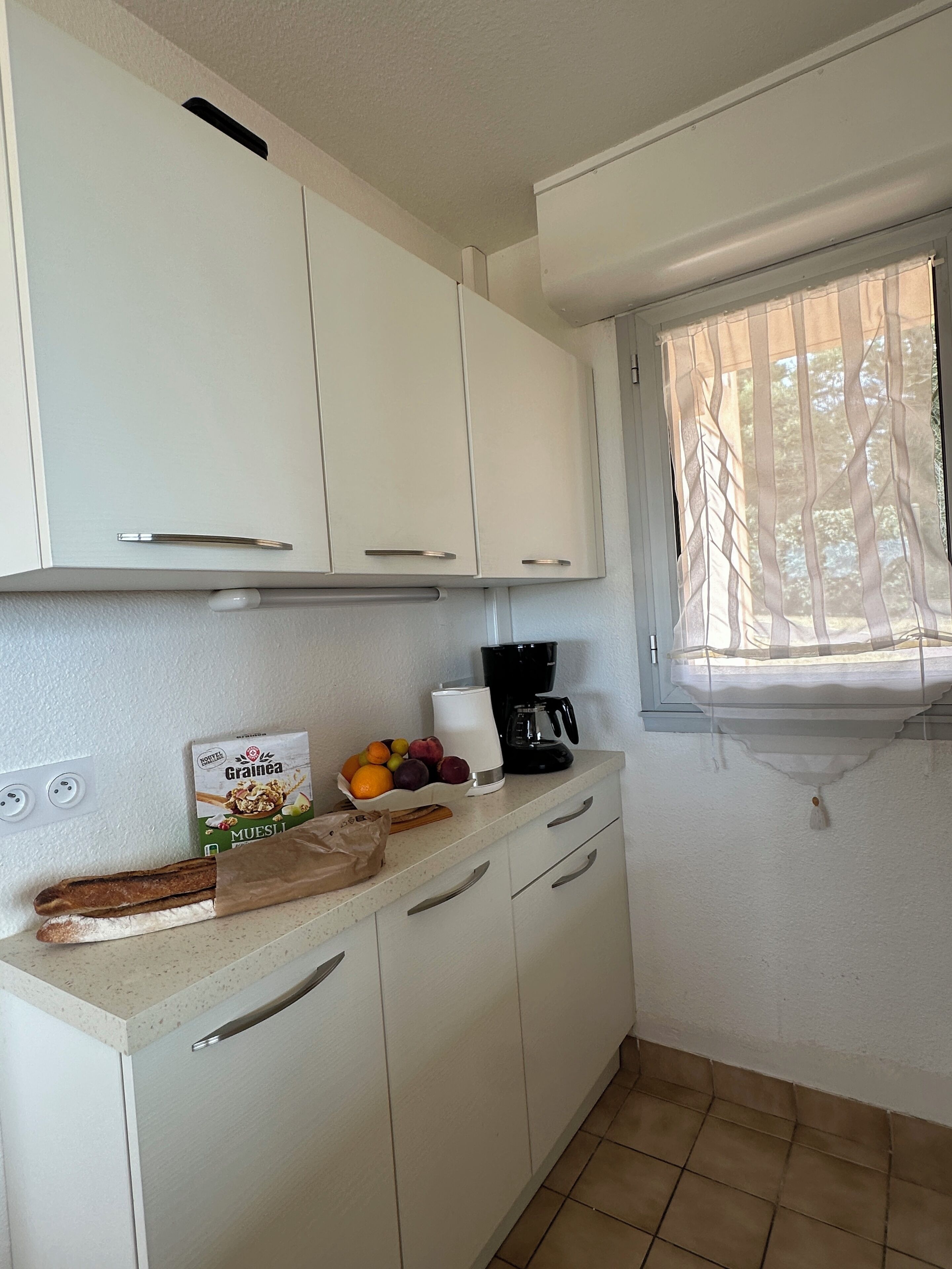 A small kitchenette with white cabinets, a coffee maker, a bowl of fruit, and a loaf of bread.