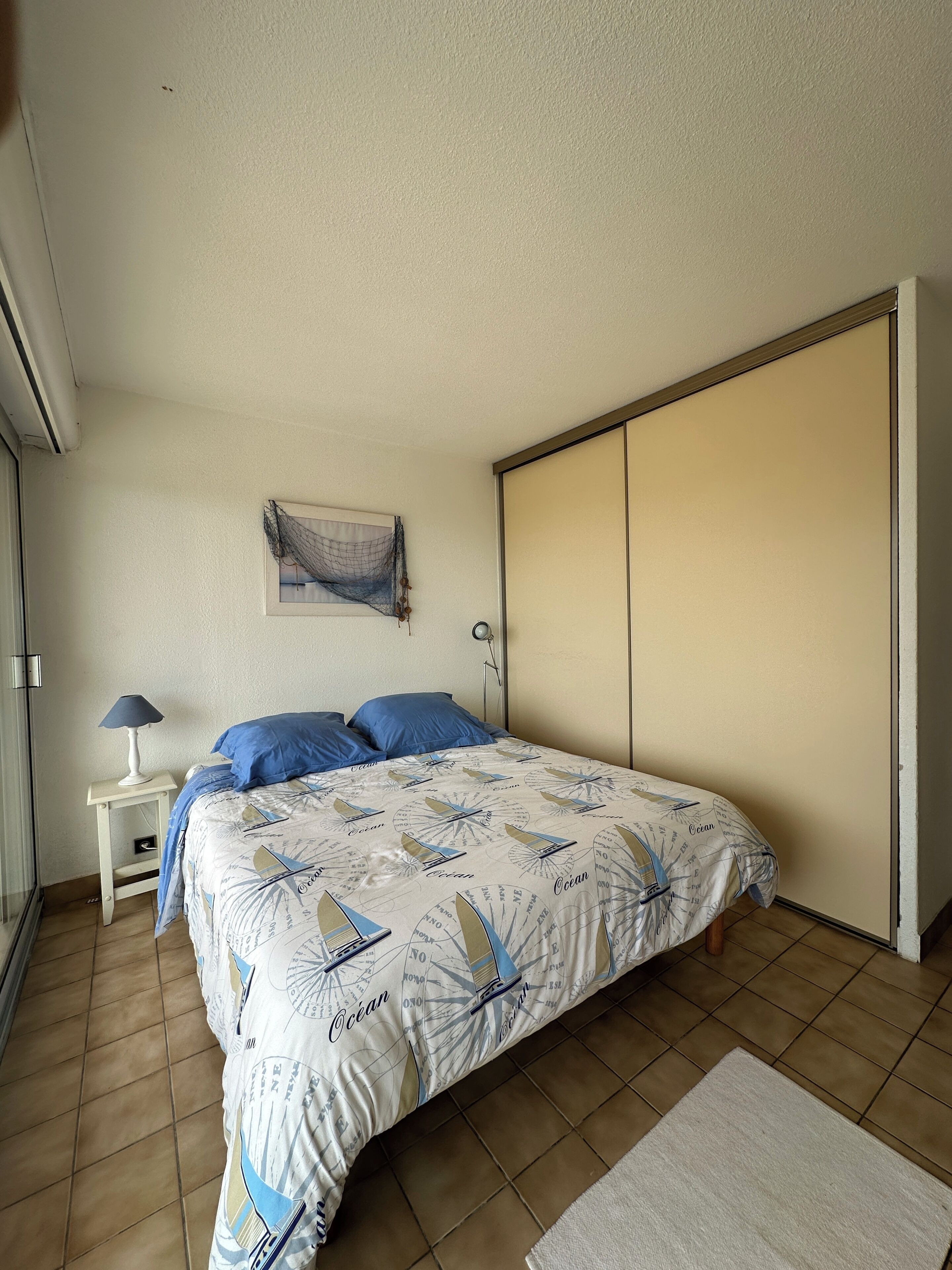 Um quarto com cama, mesa de cabeceira, luminária, obra de arte na parede e porta deslizante.