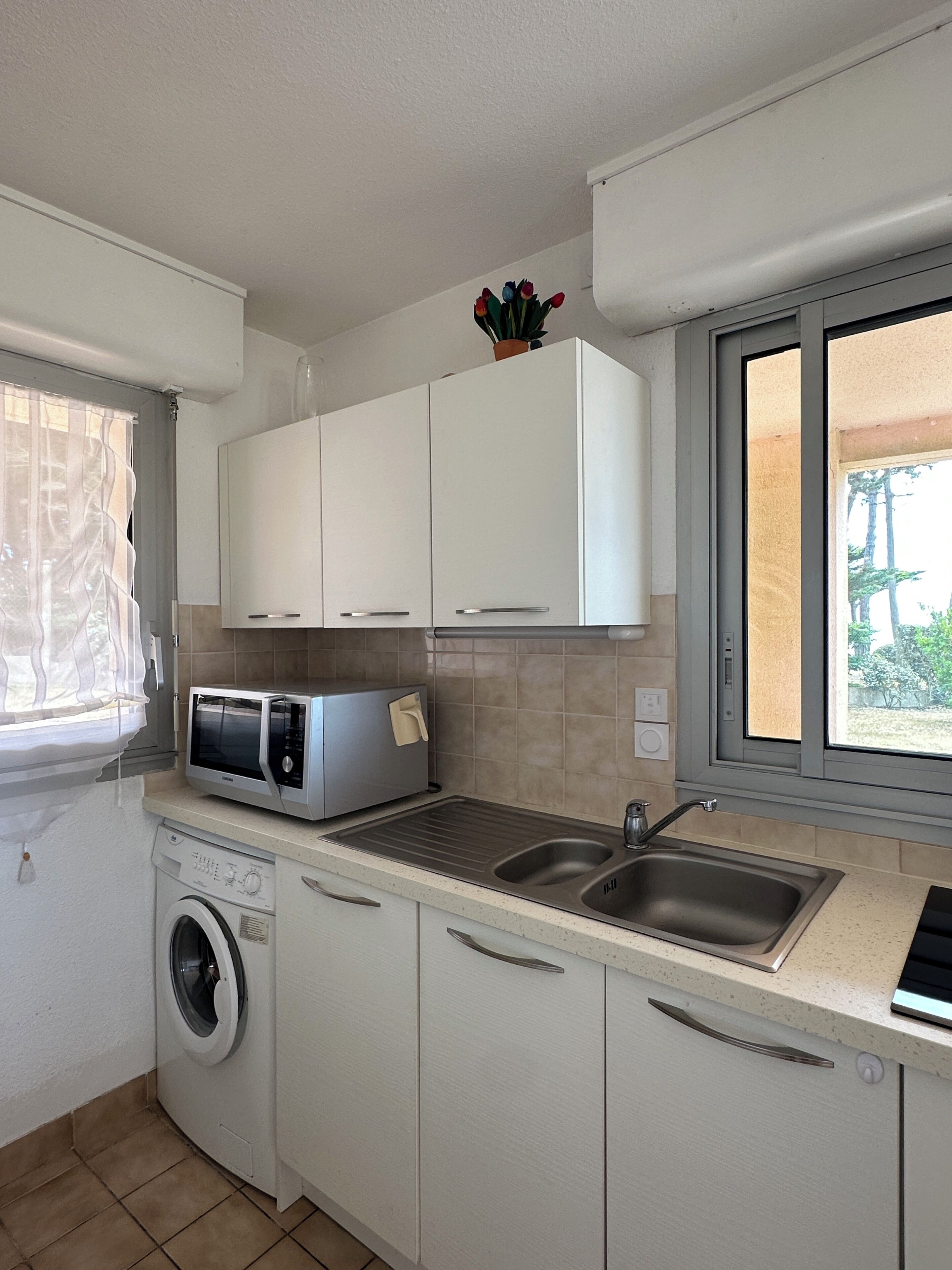 Een kleine keuken met witte kastjes, een magnetron, een wasmachine en een spoelbak.