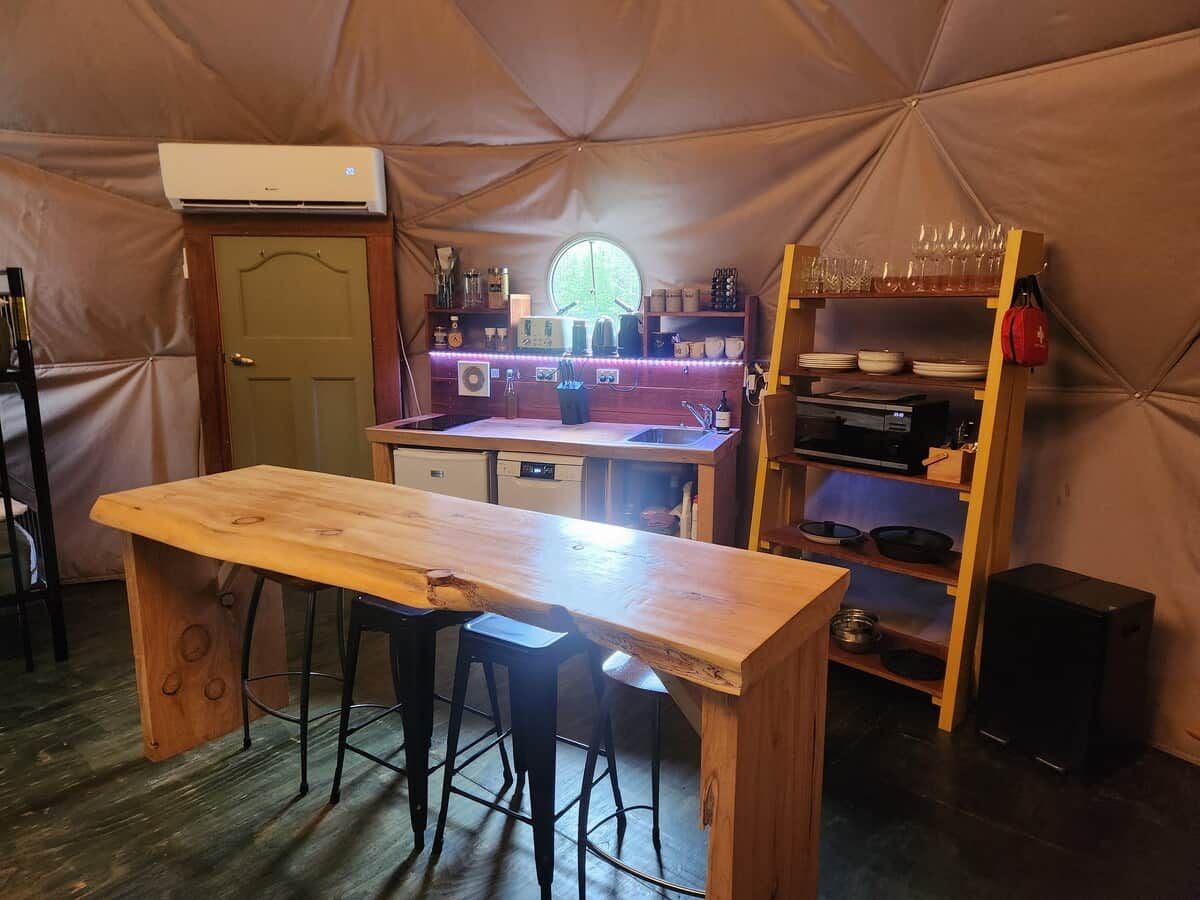 Een tent met een houten tafel en barstoelen, een keukenhoek met een spoelbak, en planken met servies en glazen.