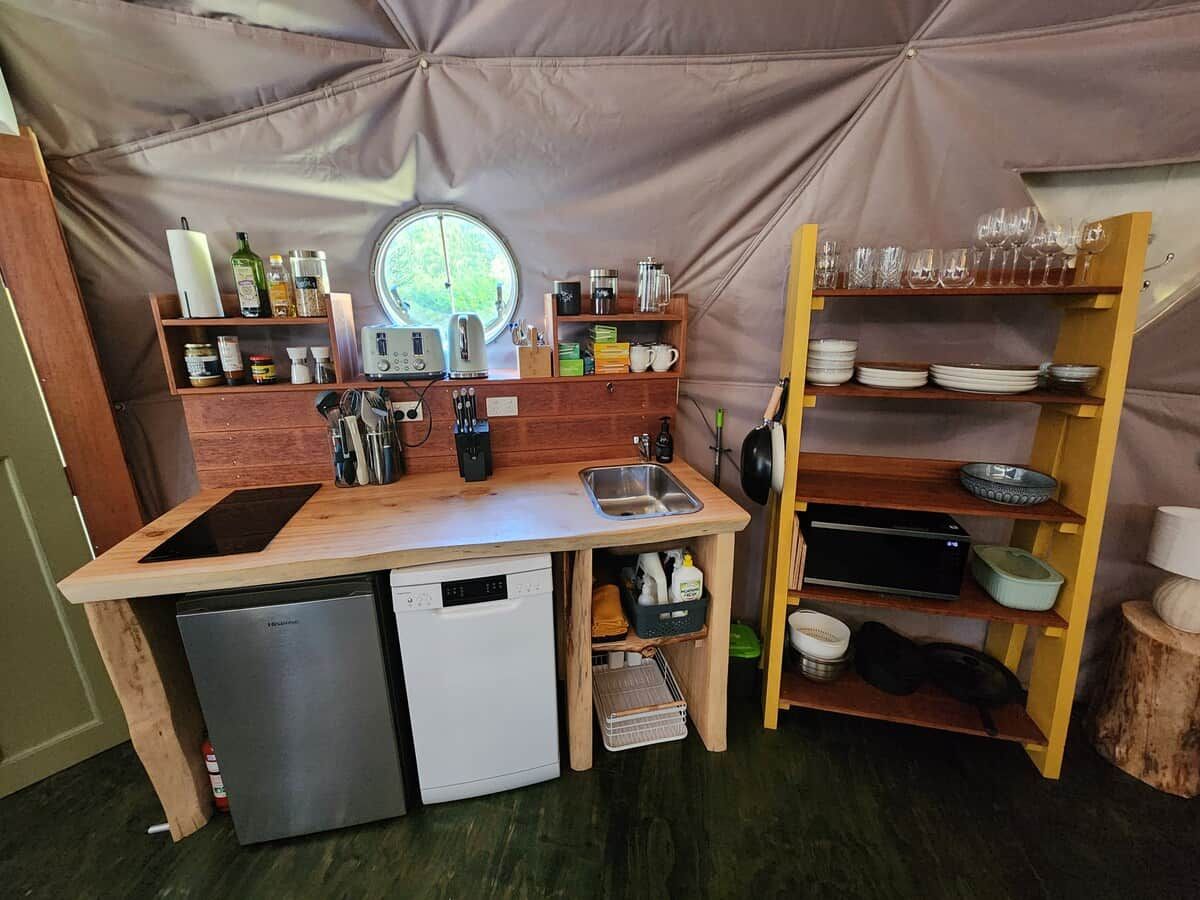Cocina compacta con lavamanos, lavavajillas y ventana. Se observan estantes con utensilios de cocina y un refrigerador pequeño.