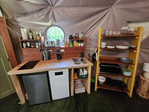 A compact kitchen with a sink, a dishwasher, and a window. There are shelves with kitchenware and a small refrigerator.