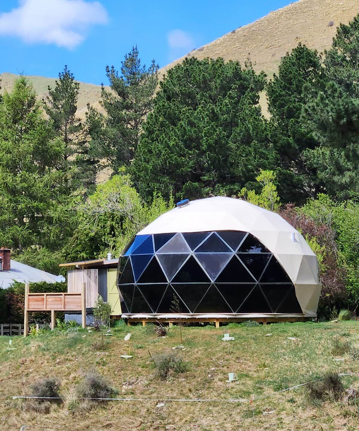 Estructura de domo geodésico en un entorno natural con una terraza de madera y un cobertizo cercano.