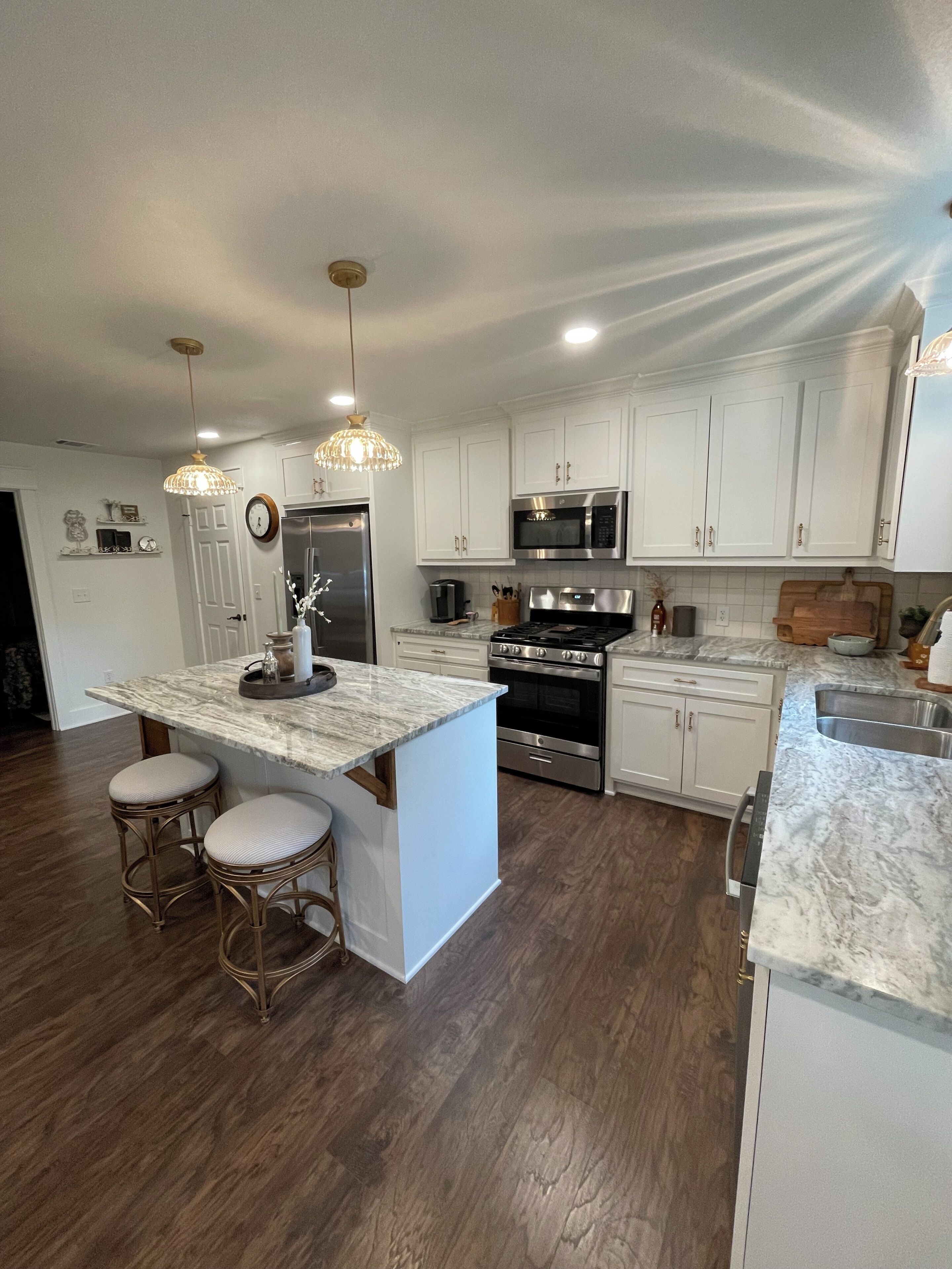 A modern kitchen with white cabinets, stainless steel appliances, a marble island with bar stools, and pendant lighting.