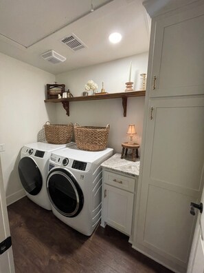 Ein Waschraum mit zwei Frontlader-Waschmaschinen, einem hölzernen Regal mit Dekoration, einem kleinen Tisch mit Lampe und einem weißen Schrank.
