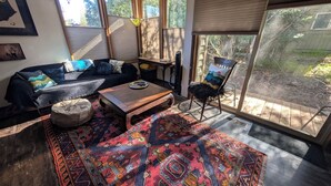 Una sala de estar con un sofá, una mesa de centro, un tapete estampado y una silla junto a un ventanal grande.