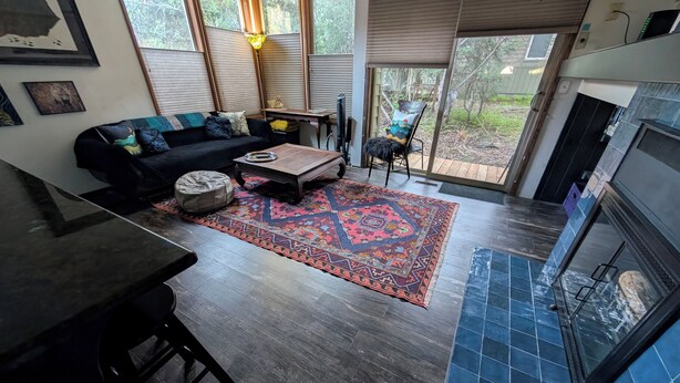 Un salón con piso de madera oscura, alfombra estampada, sofá negro, una mesita de centro y una chimenea.