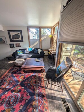 Ein gemütliches Wohnzimmer mit einer Couch, einem Hocker, einem Couchtisch, einem Teppich und einem Fenster mit Jalousien.