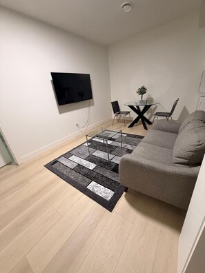 A living room with a flat-screen TV mounted on the wall, a grey sofa, a glass coffee table, two chairs, and a patterned rug. - Nice and comfortable home away from home. (Edmonton)