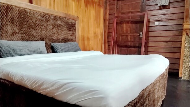 Una cama con colchón blanco y dos almohadas, cabecera de madera, puerta de madera y una mesita con una silla.