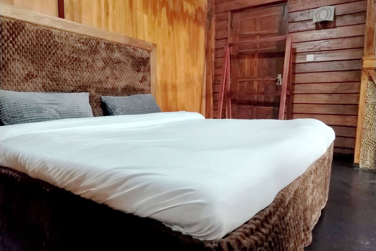 Una cama con colchón blanco y dos almohadas, cabecera de madera, puerta de madera y una mesita con una silla.
