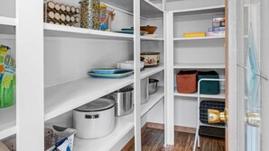 Una despensa bien organizada con estantes blancos, distintos utensilios de cocina y suelo de madera.