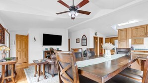 Un comedor con una gran mesa de madera, sillas, un ventilador de techo y una cocina al fondo.