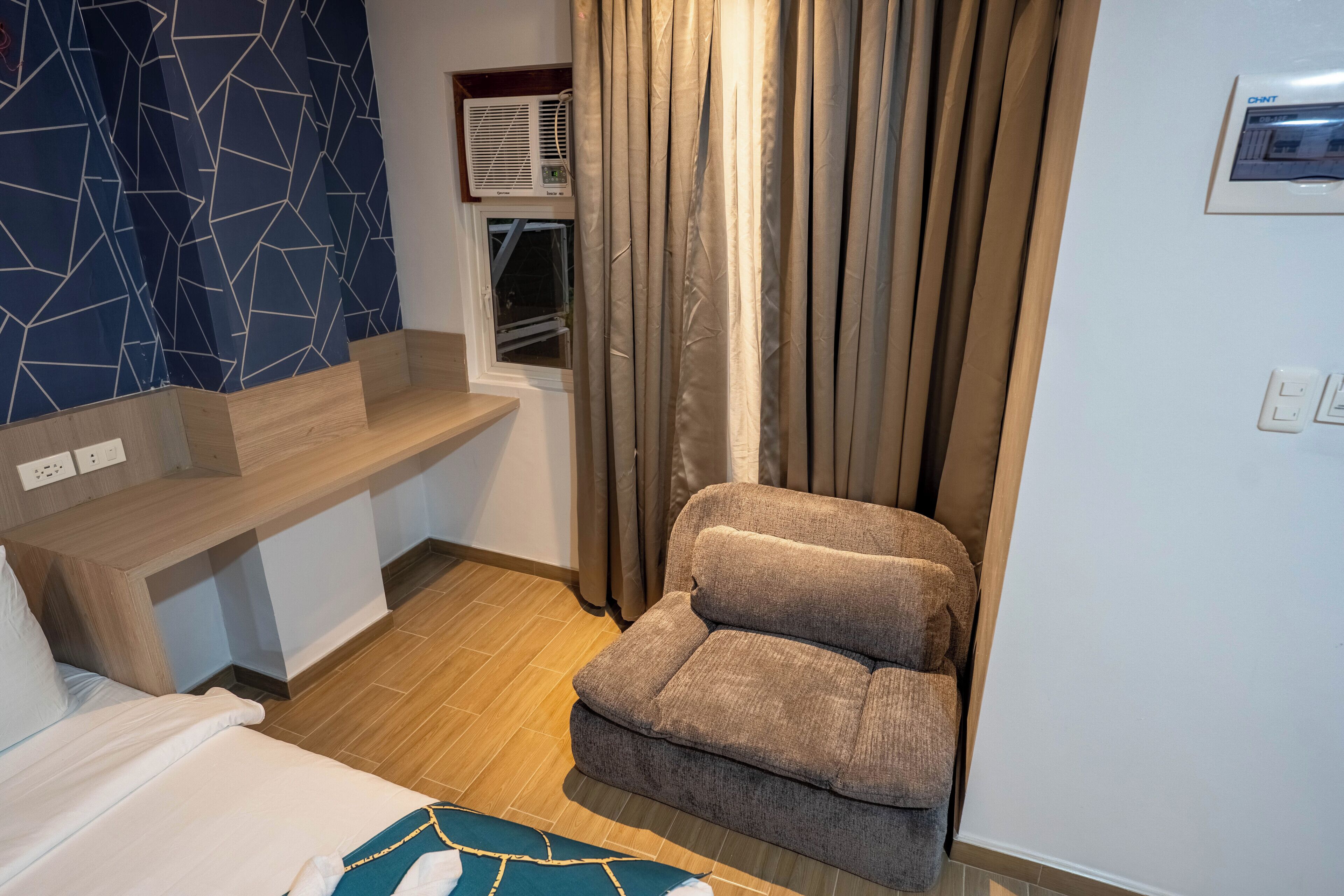 Habitación de hotel con cama, sofá, ventana con cortinas, aire acondicionado de pared y un estante pequeño.