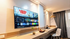 Una habitación de hotel moderna con una gran televisión de pantalla plana mostrando un programa, una mesa consola de madera, una silla blanca y una ventana con cortinas.