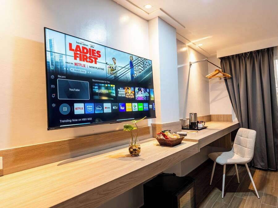 A modern hotel room with a large flat-screen TV displaying a show, a wooden console table, a white chair, and a window with curtains.
