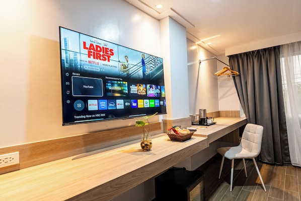A modern hotel room with a large flat-screen TV displaying a show, a wooden console table, a white chair, and a window with curtains.