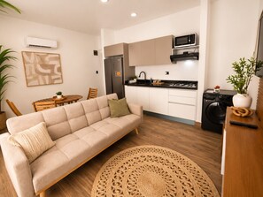 Una cocina moderna con refrigerador, microondas y lavadora. Hay un sofá color beige con un cojín estampado, un tapete redondo tejido y una mesa de comedor con sillas.