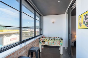 A room with a foosball table labeled 'SOCCER,' a window with a view of buildings, and a wall-mounted light fixture. - Vibrant 3BR Petone Stay w Foosball & Harbour Views (Lower Hutt)
