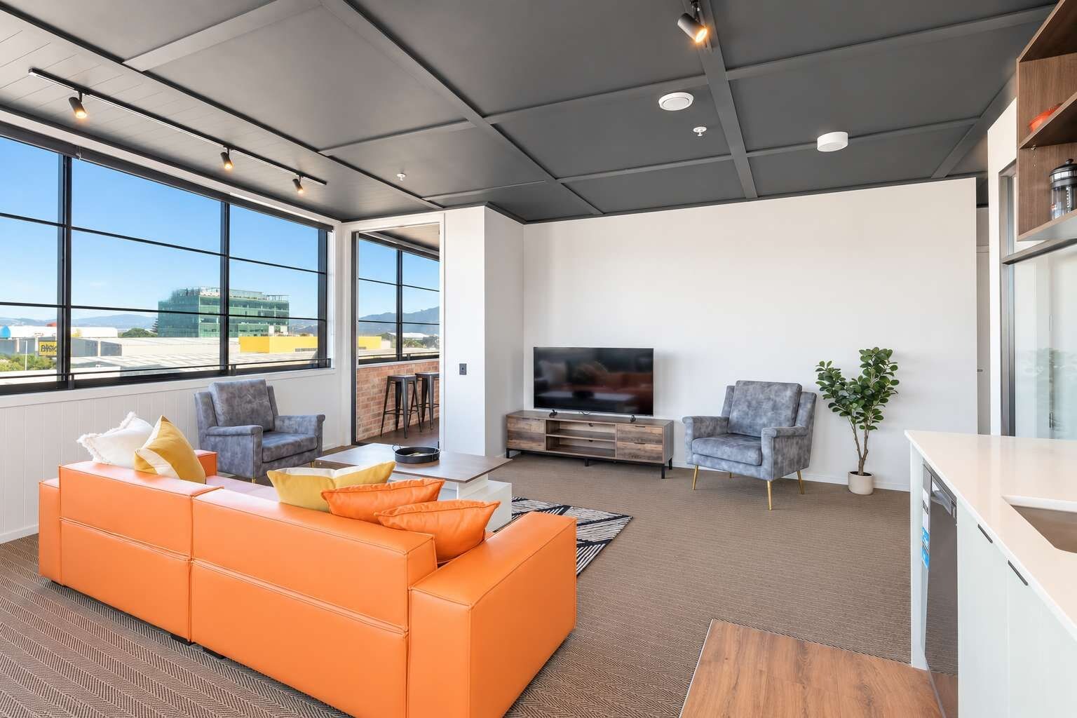 A modern living room with a large orange sofa, grey armchairs, a flat-screen TV, and a dining area with a bar.