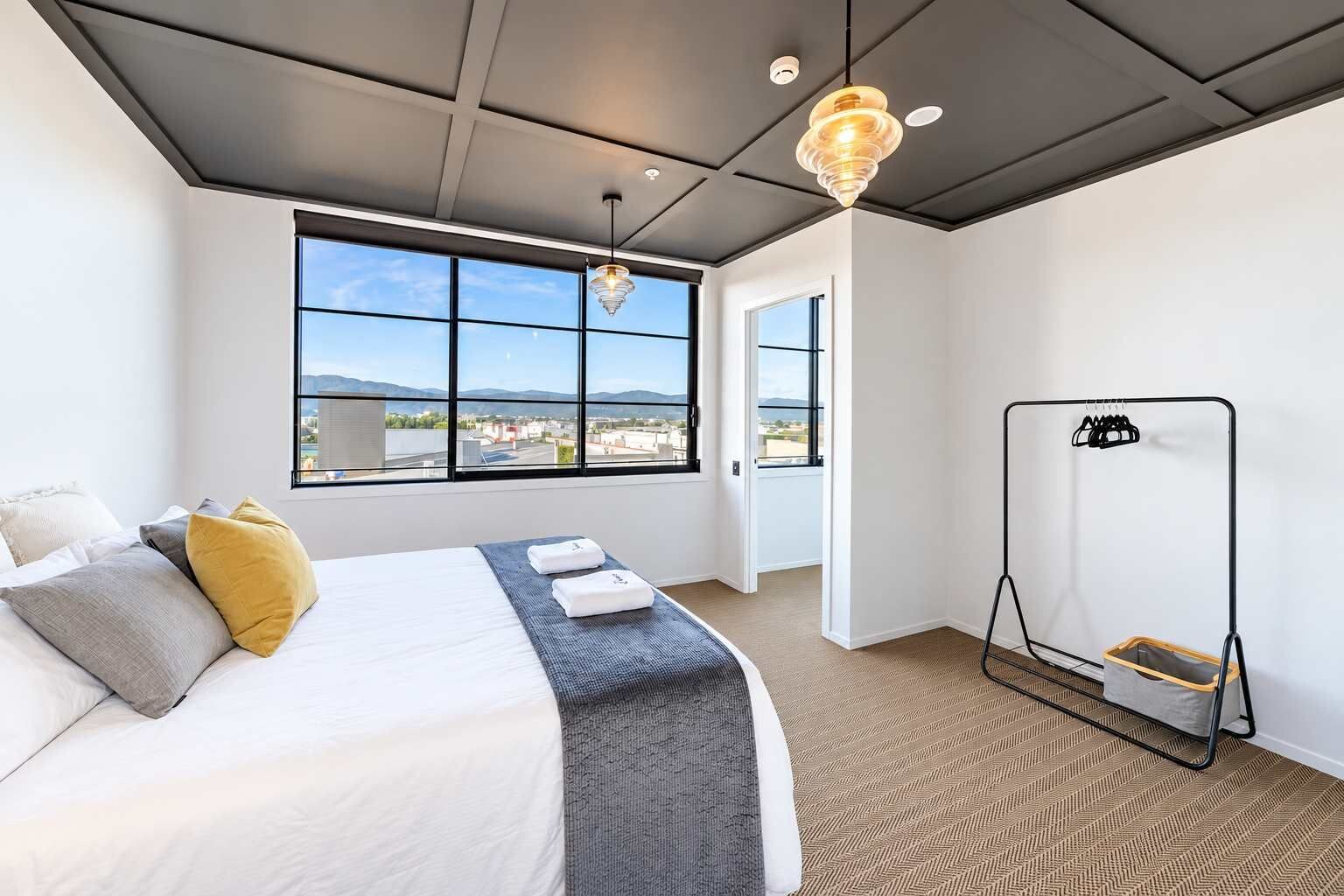A hotel room with a large bed, a view through the window, and a metal rack with towels.