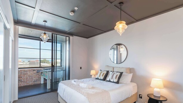 Une chambre d’hôtel avec un grand lit, un miroir rond, deux lampes de chevet, une fenêtre avec vue et un balcon équipé d’un baby-foot.