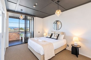 Une chambre d’hôtel avec un lit, des lampes de chevet, un miroir et une vue sur une table de baby-foot, visible à travers une porte vitrée.
