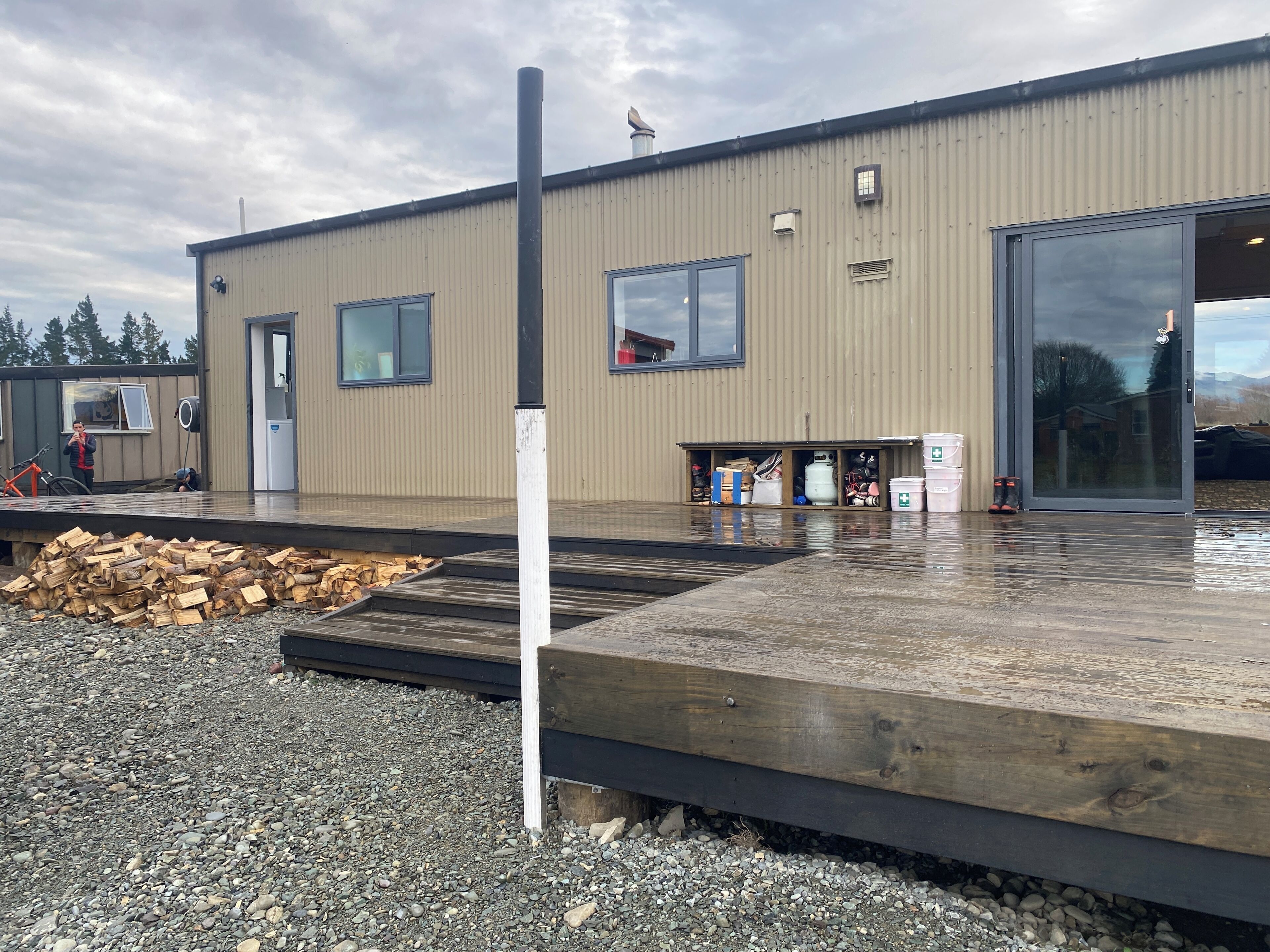 A building with a large garage door, a wooden deck, and a gravel area with a pile of wood.