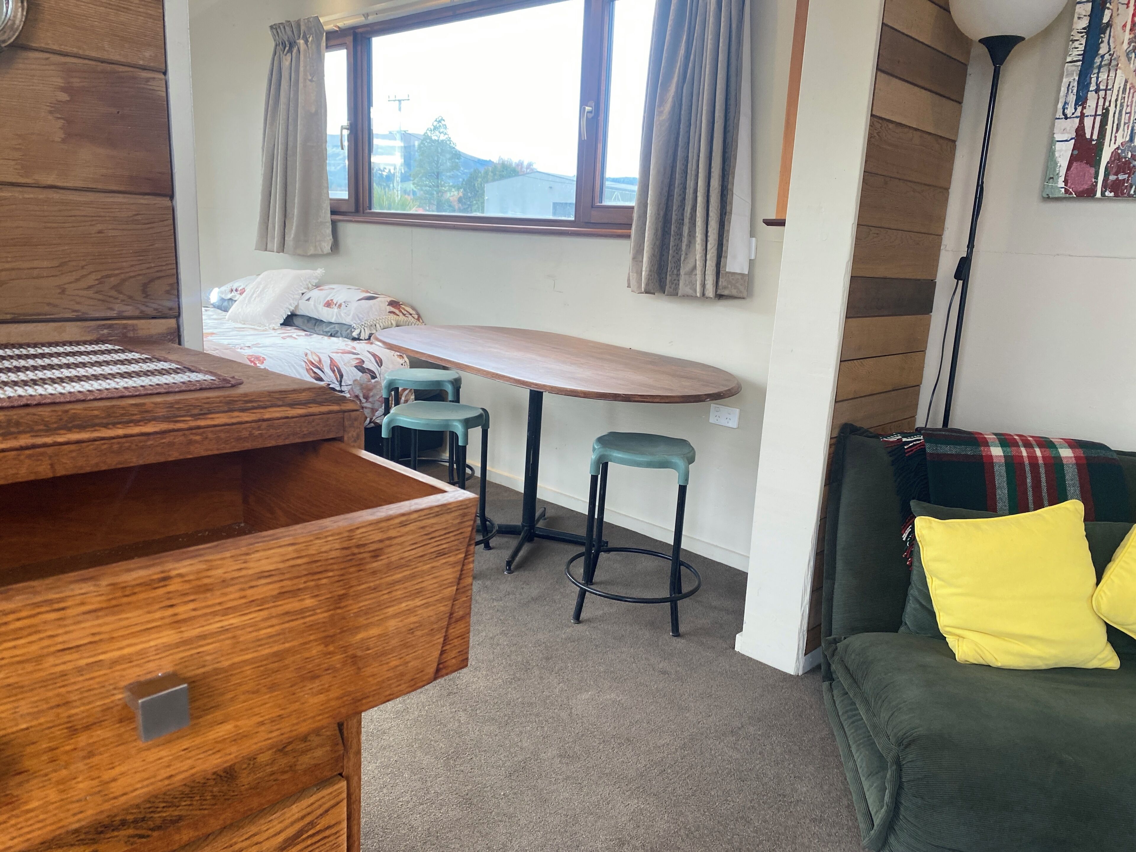 Ein gemütliches Zimmer mit einem Holztisch, zwei Hocker, einem Bett mit floralem Bettbezug, einem Fenster mit Vorhängen, einem grünen Sofa mit gelbem Kissen und einer Wandlampe.