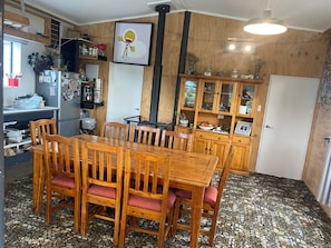 A dining area with a wooden table and chairs, a kitchen with a refrigerator and shelves, and a cabinet with glass doors.