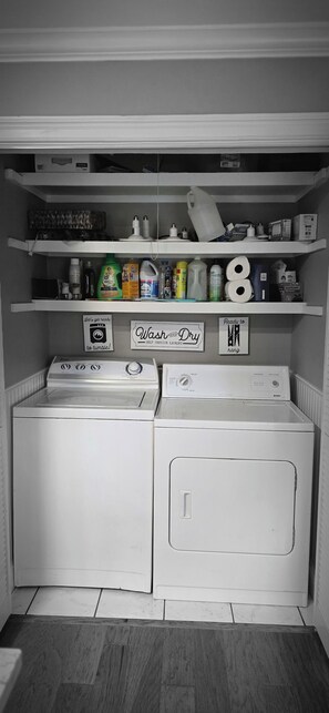 A laundry area with two stacked washers and dryers, shelves with cleaning supplies, and signs indicating self-service laundry options. - Midtown Oasis 
3 Bedrooms house in  Rogers with  AC, private pool and more (Rogers)